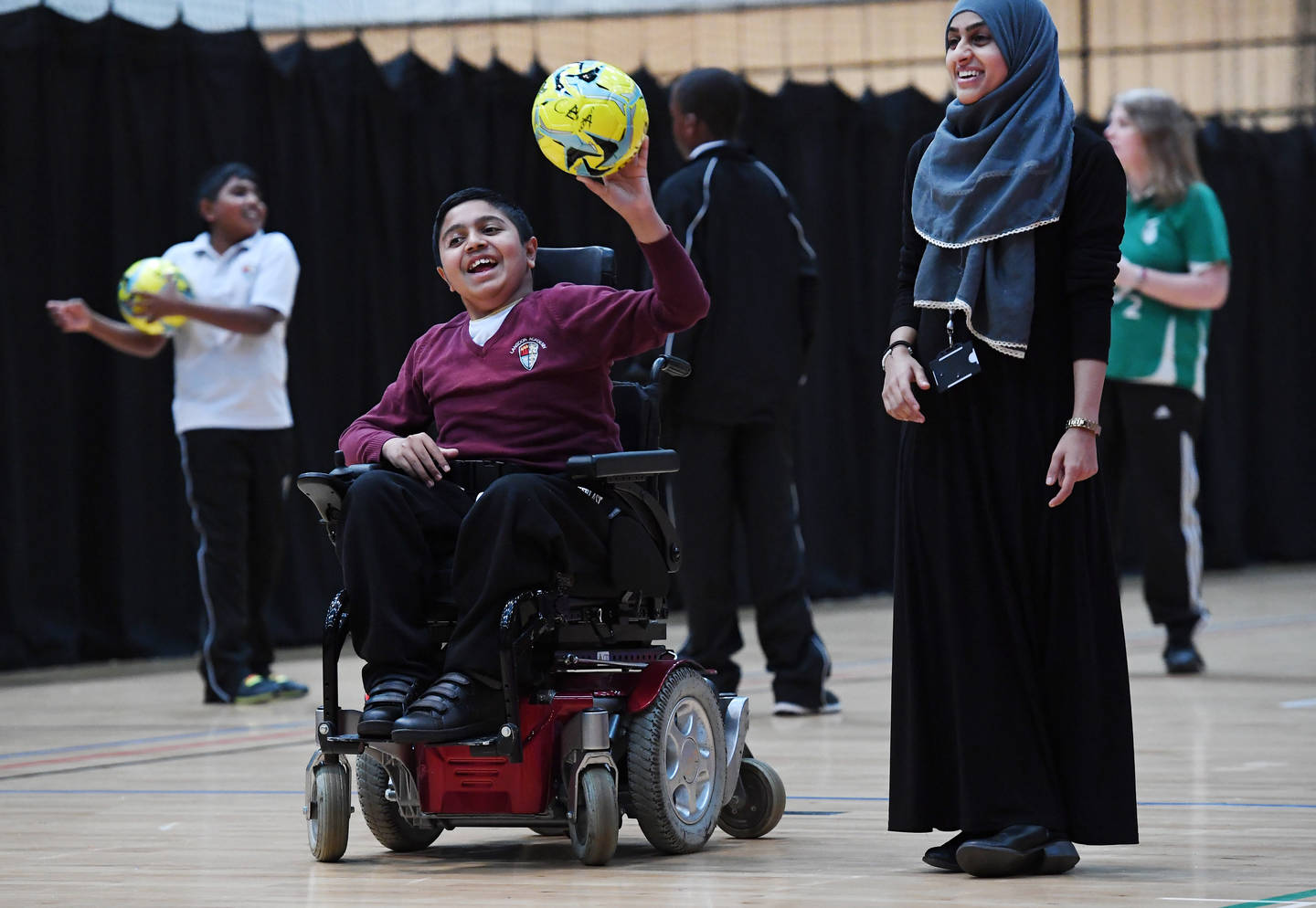Boy in powerchair with football in hand laughing with girl stood next to him smiling.