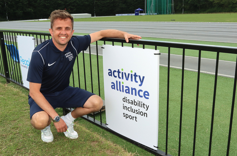 Adam Blaze, CEO of Activity Alliance, crouches next to the Activity Alliance logo on a board.