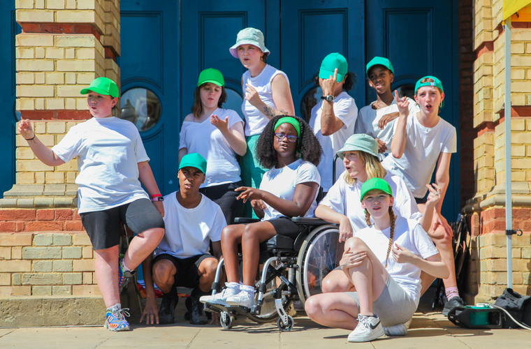 A group of children in white t-shirts and green caps stand in front of a blue door looking on.