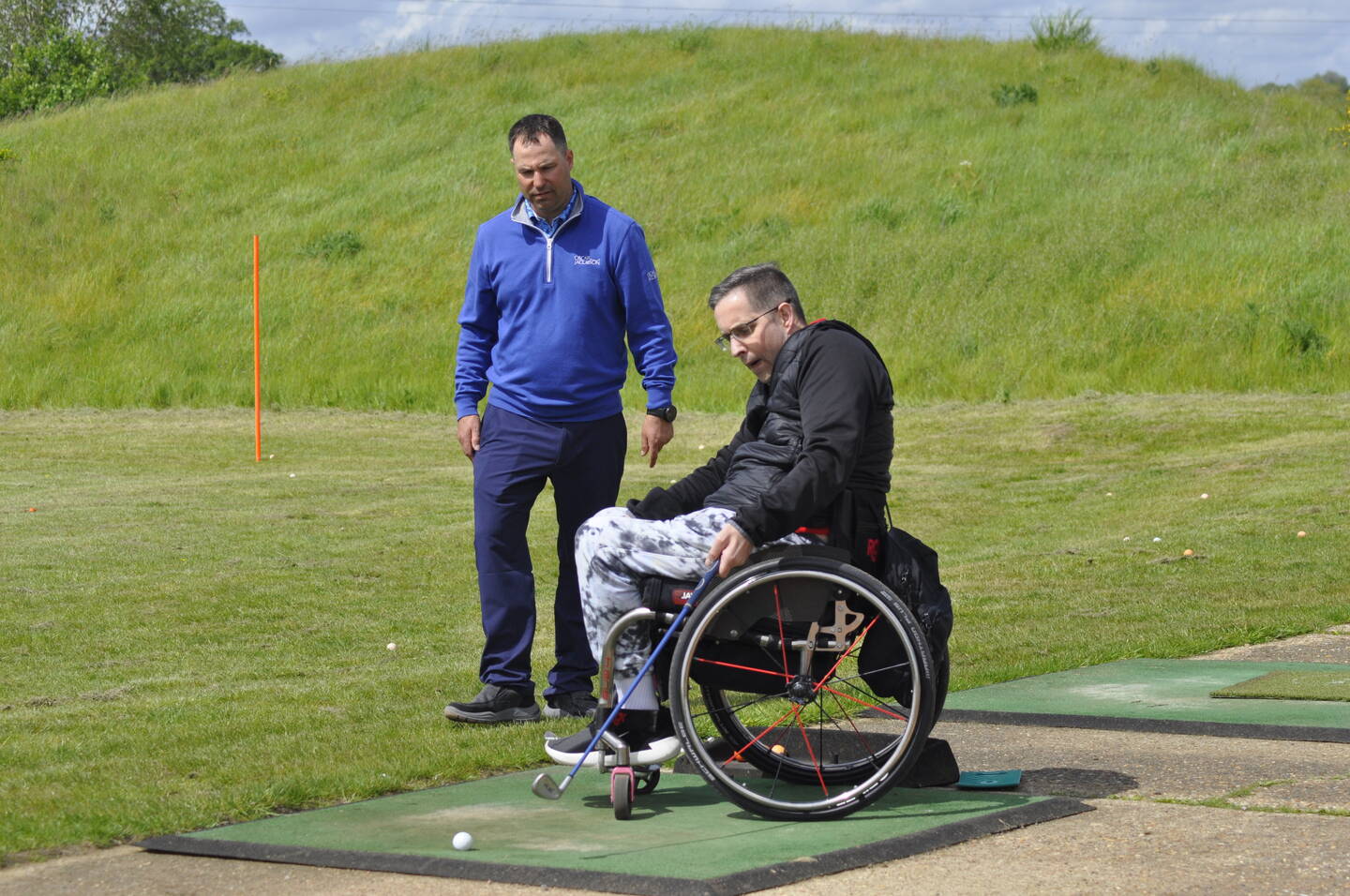 A disabled man in a wheelchair is playing golf. He is holding a golf club, ready to hit a golf ball on a mat. There is a coach behind him watching his moves. They are on a golf course.