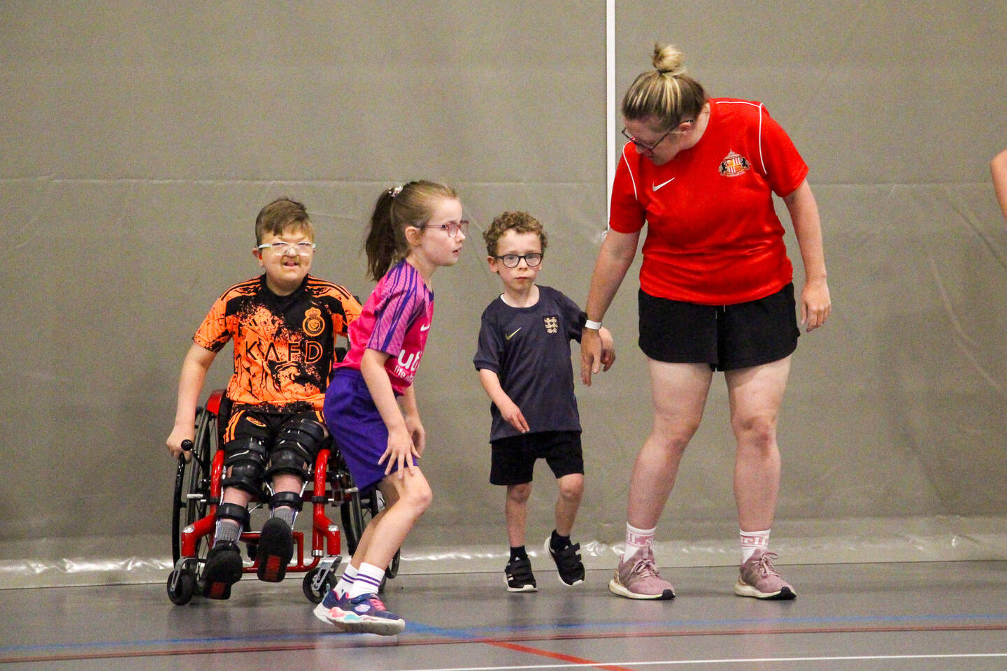 A sports coach runs holding hands with a child at a multi-sport session. There is also a group of children involved including a child using a wheelchair.