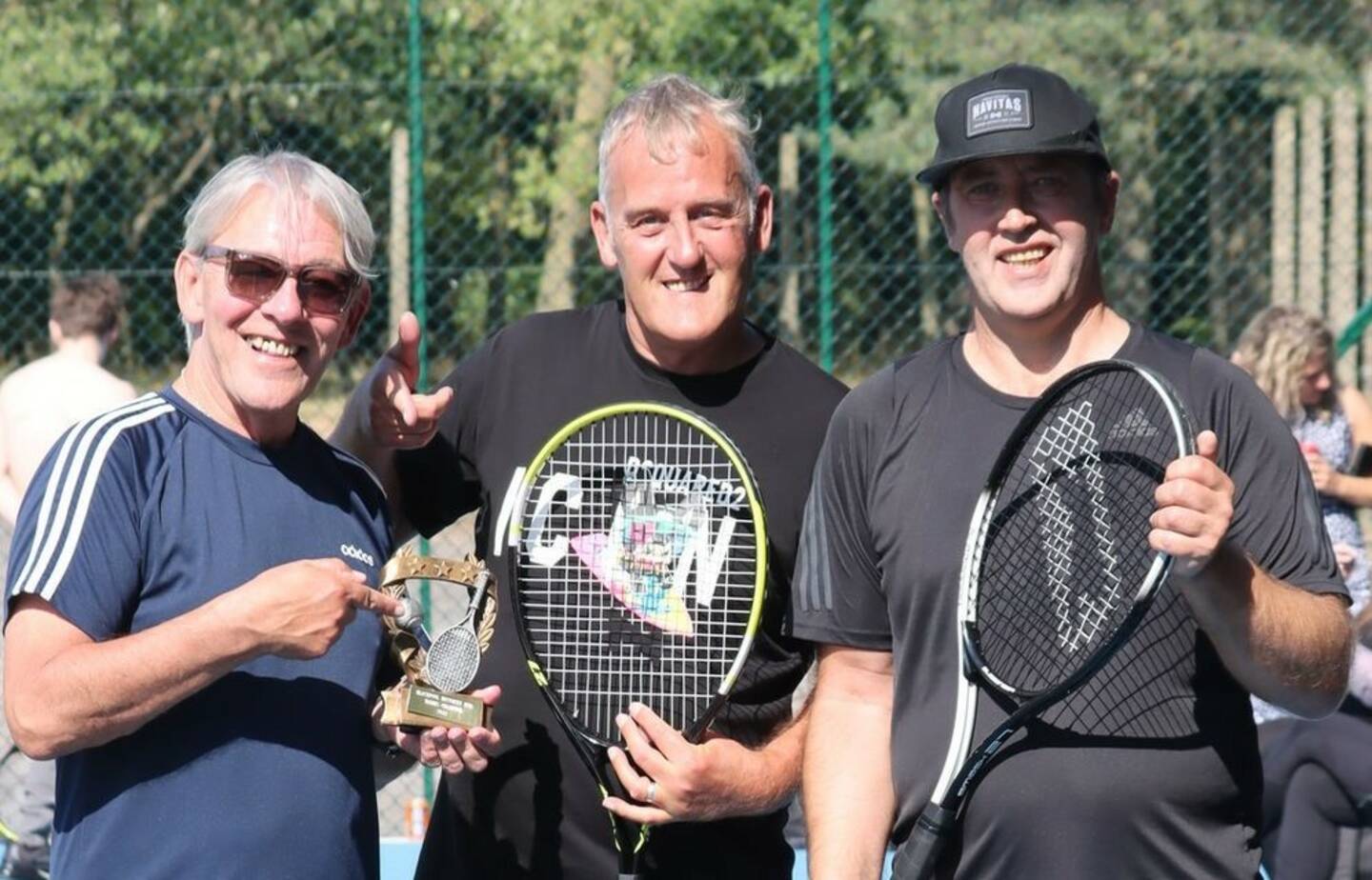 Three men smile holding tennis racquets and a trophy.