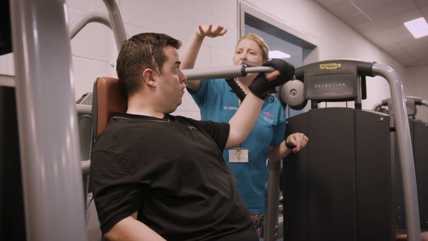 A female trainer helps a man on a seated weights machine.