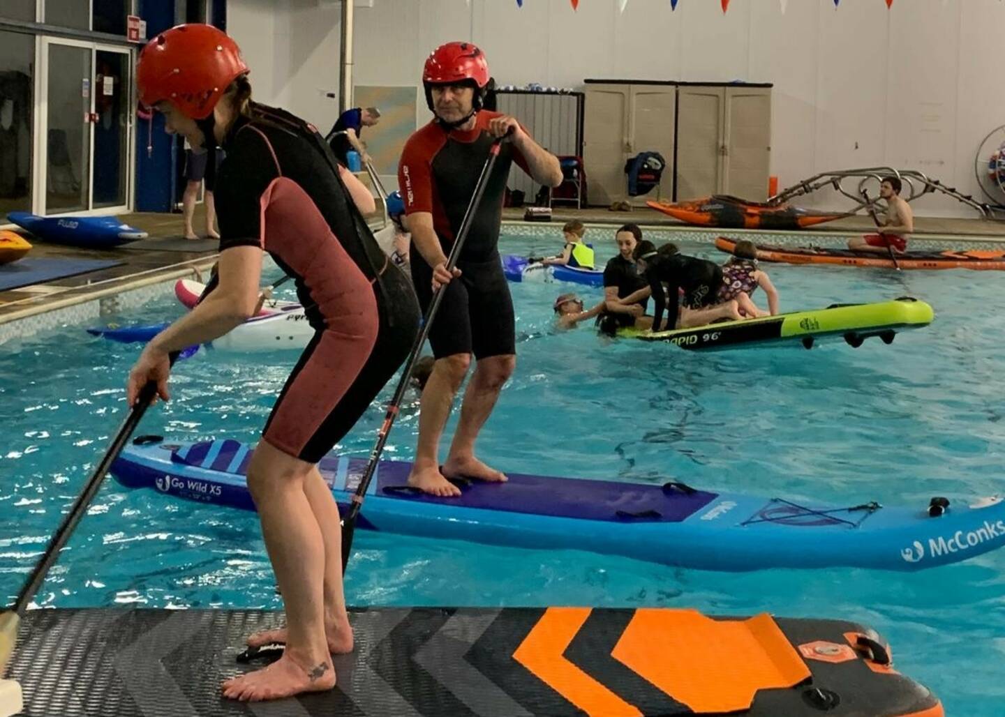 Two people stand on paddleboards in a swimming pool. A group of children play behind them.