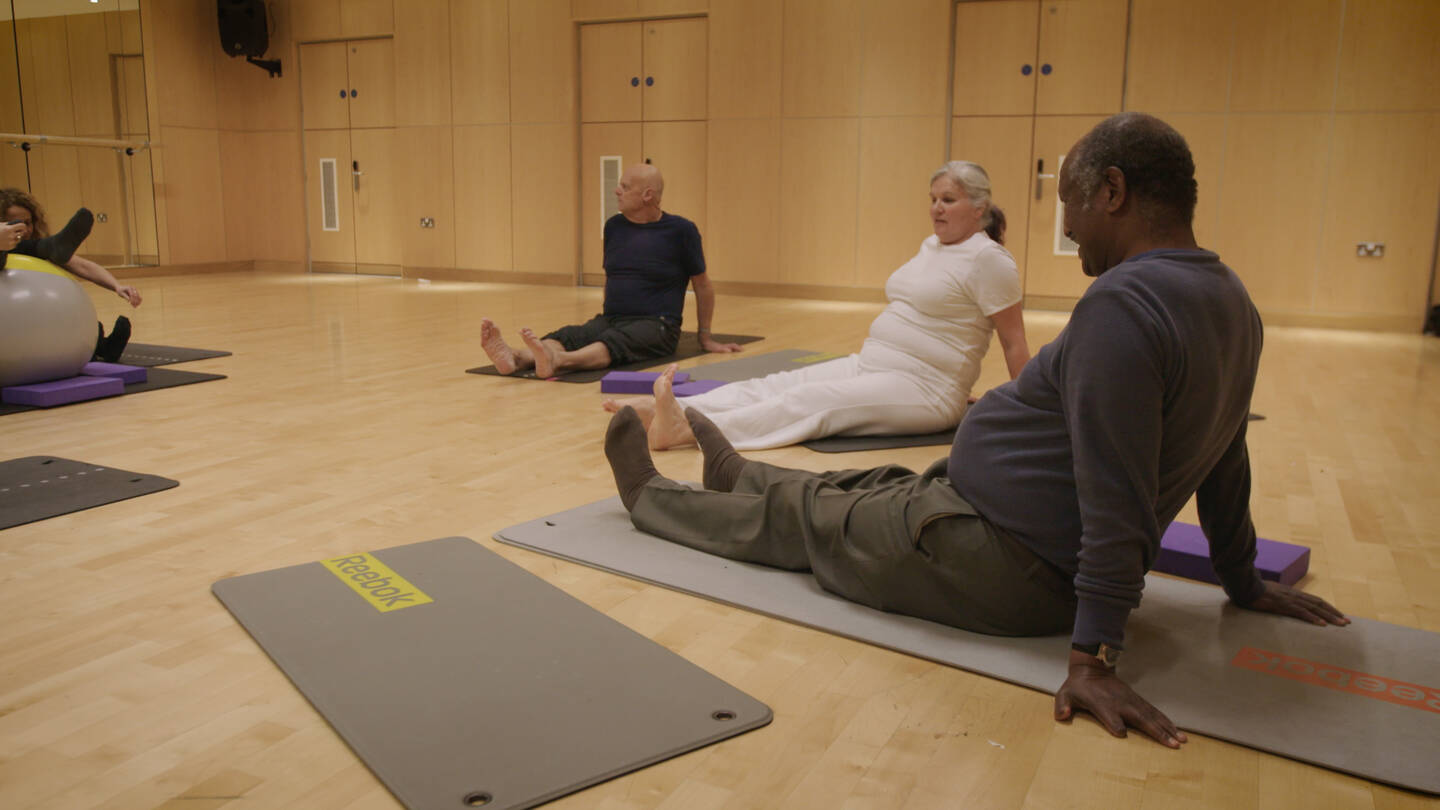 Two men and a woman sit on yoga mats in a gym room with a mirror on the wall.